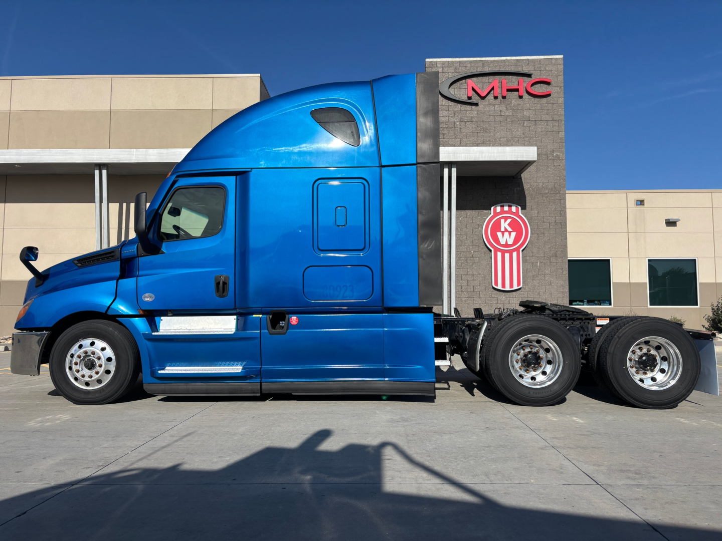 Truck side profile showing a blue Freightliner PT sleeper cab with a black chassis and three rear axles, KW emblem on the sleeper, dealership building in background.