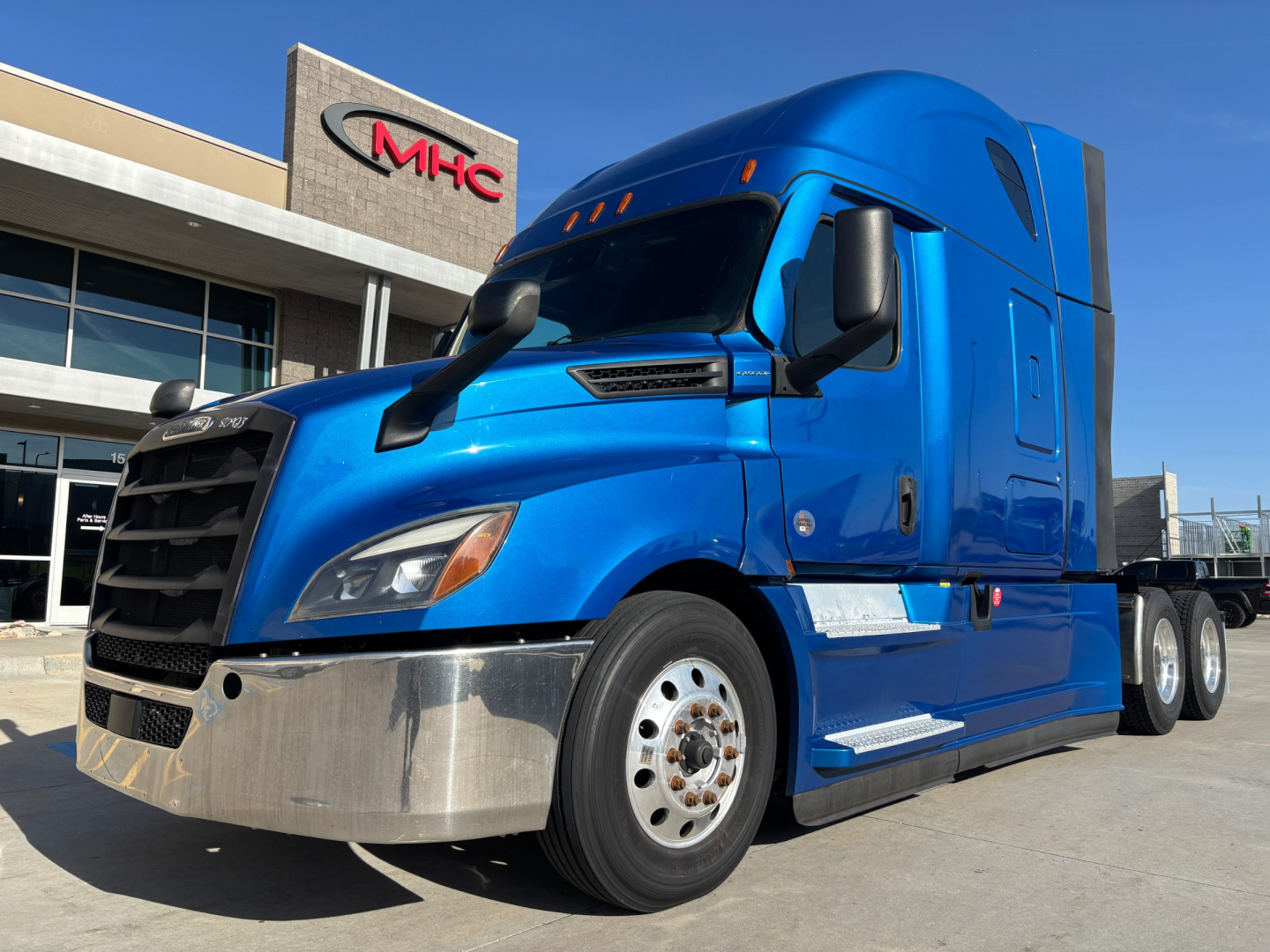 Freightliner semi truck in blue exterior parked outside dealership showing chrome front bumper and aerodynamic sleeper cab.