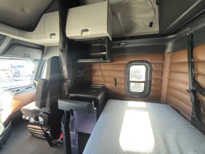 Truck cab interior with bunk bed area, overhead shelf, storage cabinet, and small window. Passenger seat visible, brown padded wall, light coming through window.