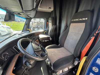 Truck cab interior showing steering wheel and Cascadia seat. Dashboard with gauges, driver seat with label Cascadia, window and side mirror view.