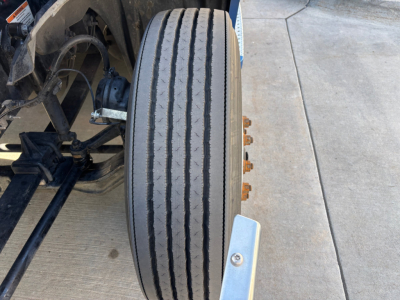Front wheel view of a 2023 Freightliner PT126064ST conventional sleeper truck showing single tire with dual grooves and adjacent suspension components.