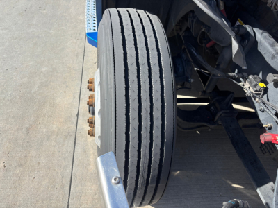 Truck front wheel and suspension view showing dual rear tires and axle components under a Freightliner PT126064ST cab with visible bolts.
