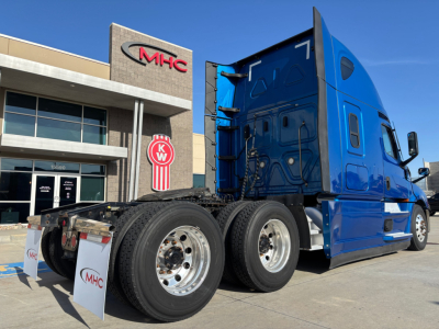 Freightliner PT126064ST sleeper tractor showing blue cab and dual rear axles with chrome wheels in front of a dealership.