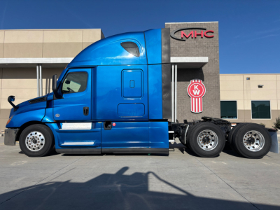 Truck side profile showing a blue Freightliner PT sleeper cab with a black chassis and three rear axles, KW emblem on the sleeper, dealership building in background.