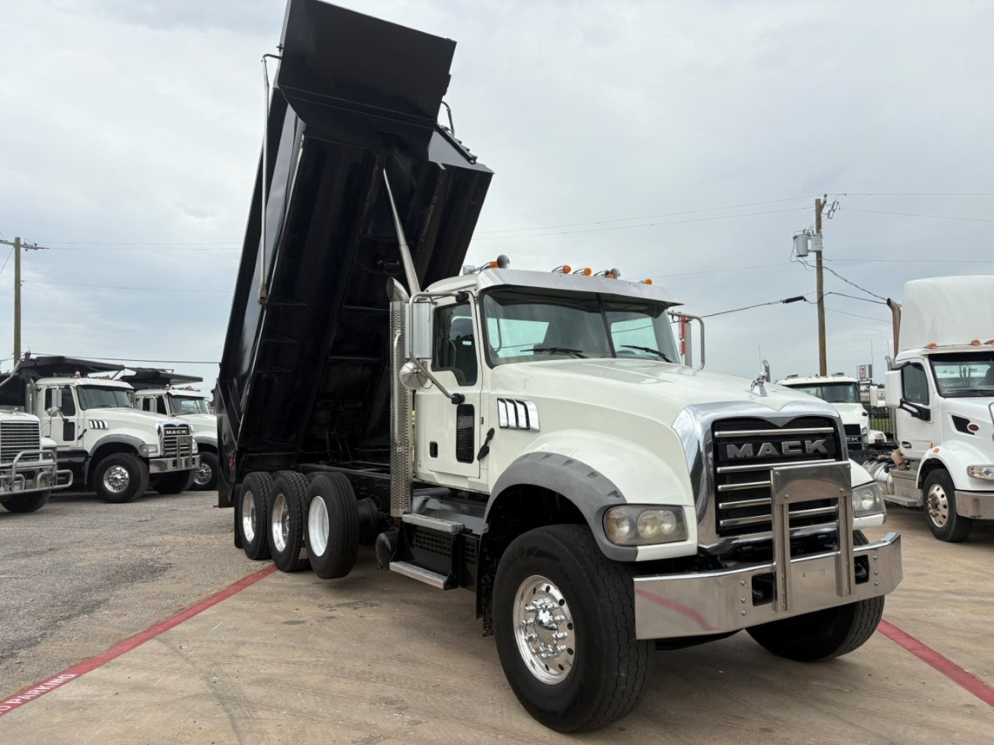 Mack GR64F dump truck with raised dump body in a lot; front chrome bumper, grille bearing 'MACK' emblem, multiple axles, white cab, side mirrors, and yellow clearance lights.