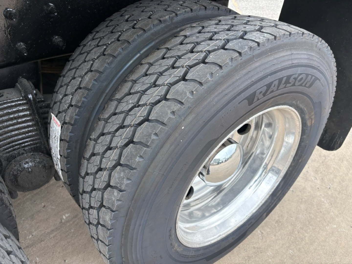 Truck wheel and tire close-up on a 2020 Mack GR64F dump truck showing a Ravison/RA… tire with tread pattern and shiny chrome rim; part of rear axle and suspension visible.