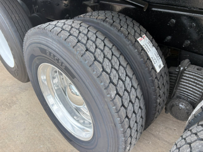Dual rear truck tires with visible tread and white wheel rims. A label is attached to the nearby suspension area, with partial text on the tire sidewall reading 'RACE' and factory components nearby.