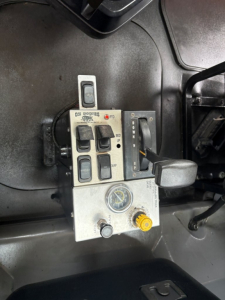 Control panel on Mack GR64F dashboard showing multiple switches, a gauge, and a yellow knob; features include a rectangular switch cluster with labels and a black lever, and a round gauge with a yellow knob nearby.