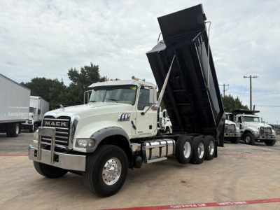 Mack GR64F dump truck in-tilt position showing front grille with Mack lettering and dual rear wheels, white cab, black dump body raised, dealership lot background.