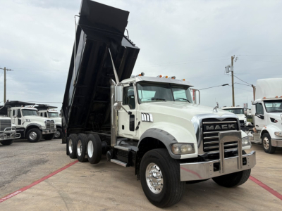 Mack GR64F dump truck with raised dump body in a lot; front chrome bumper, grille bearing 'MACK' emblem, multiple axles, white cab, side mirrors, and yellow clearance lights.