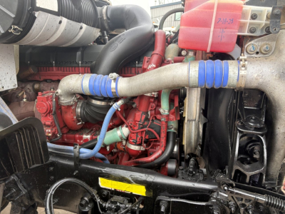 Engine bay of a 2020 Mack GR64F dump truck showing red engine block, blue silicone hoses, black radiator hoses, plastic coolant reservoir, and metal piping.