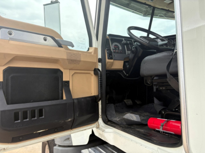 Truck cab interior visible through open door; dash, steering wheel, pedals; beige/black interior; red fire extinguisher on floor; exterior tan body panels near door hinge; VIN/stock info likely on page.