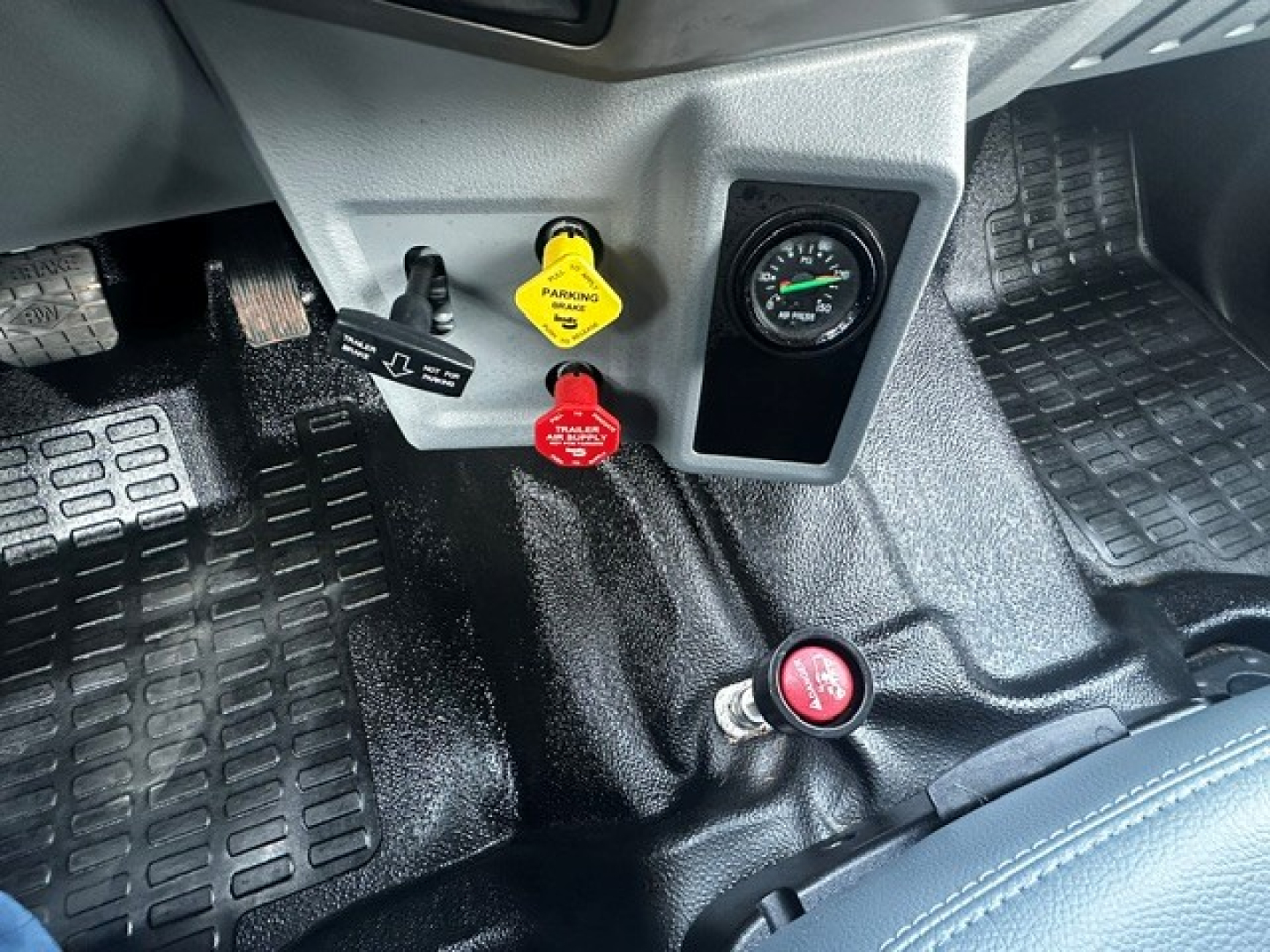 Dashboard area of a 2025 Ford F750 with ignition key and two colored dials near the center console; visible labels include a yellow 'PARKING BRAKE' handle and a red 'FUEL' cap/knob; textured rubber floor mats and pedals are visible.