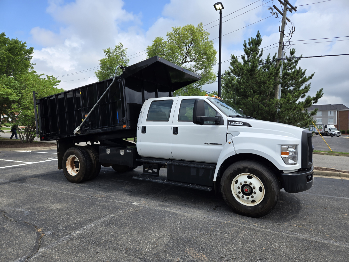 White 2025 Ford F750 dump truck with black dump bed in a parking lot, side view showing cab and front wheel with Ford badge.