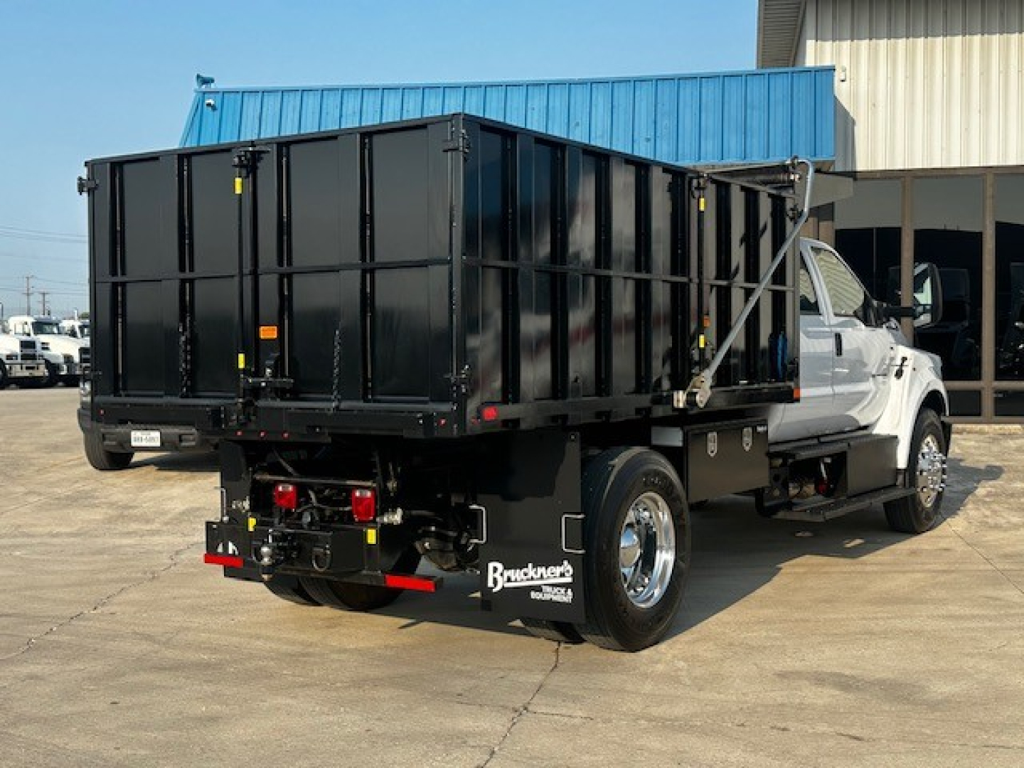 Dump truck chassis with a black dump bed on a white Ford F-750 cab and chassis; rear view showing hitch, taillights, and hydraulic dump mechanism.