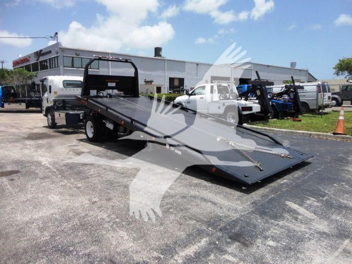 Isuzu NRR rollback truck with a tilted flatbed/wheel-lift, showing a long black steel ramp surface and side rails; background includes buildings and other trucks.