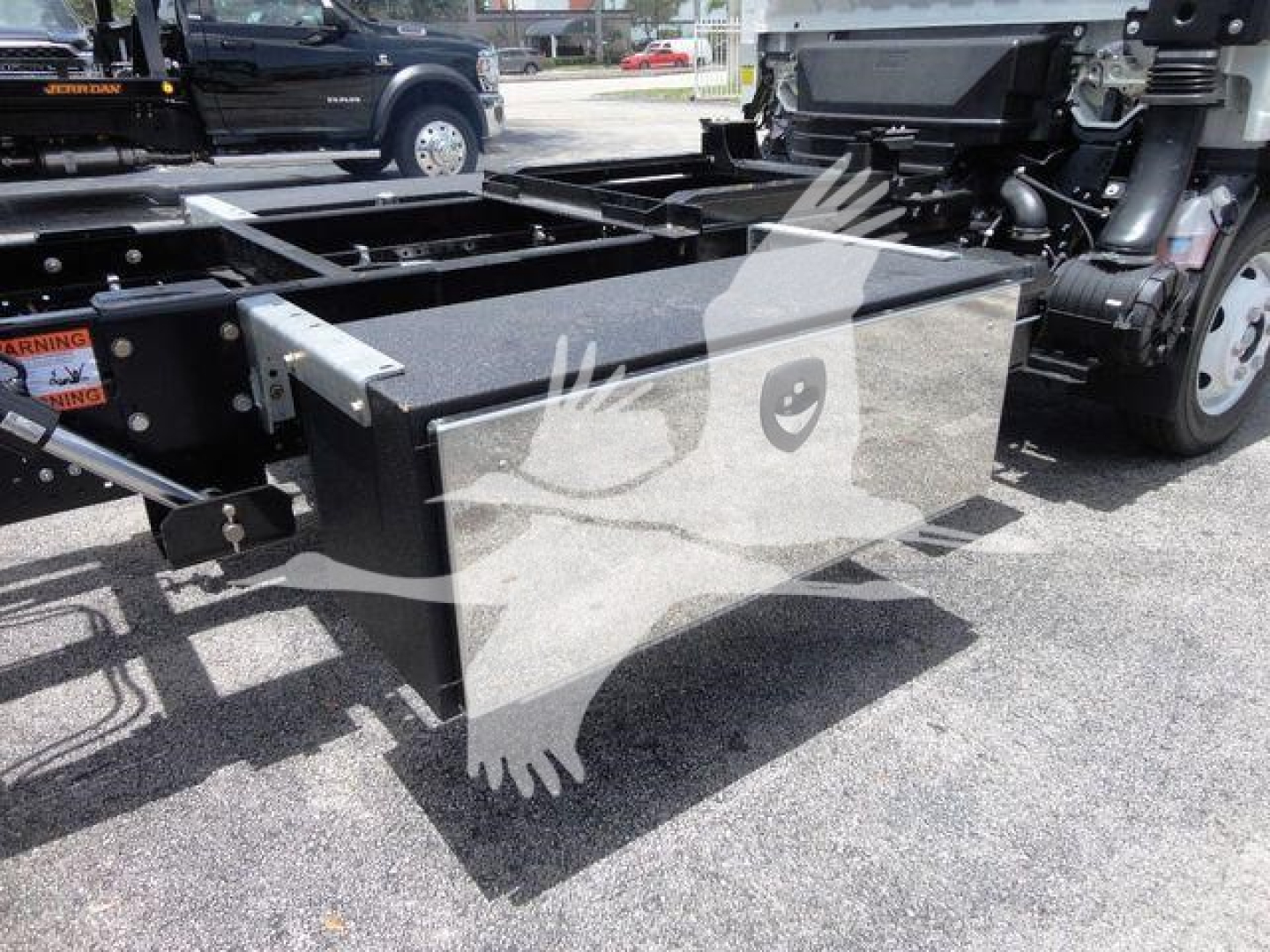 Isuzu NRR rollback truck chassis with a large rectangular metal toolbox mounted between the frame rails; visible components include a black toolbox, frame rails, and part of the cab suspension.