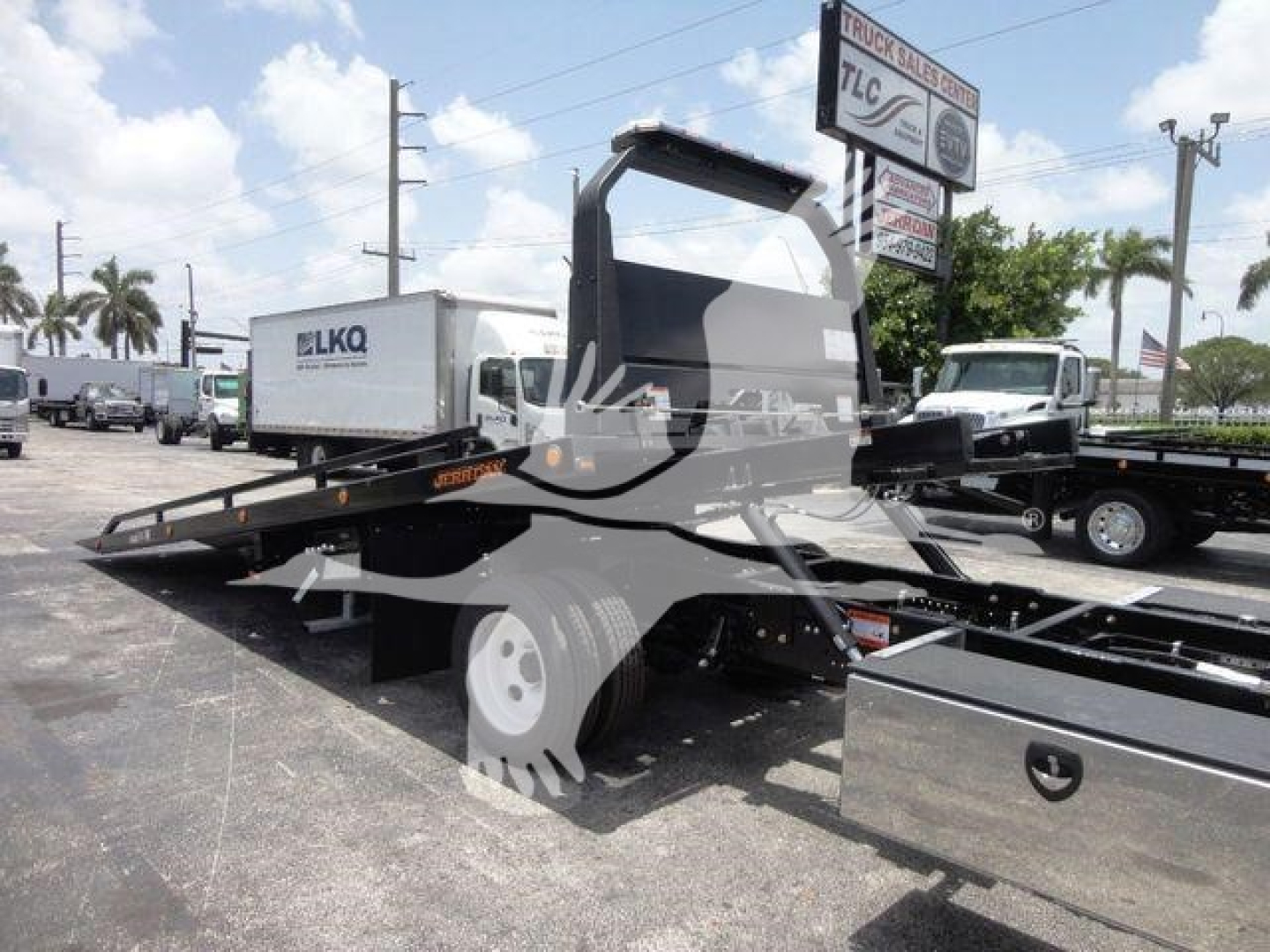 Isuzu NPRR rollback tow truck side view showing hydraulic tilt bed and cab with open-frame crane, chrome wheel, and nearby equipment in a lot.
