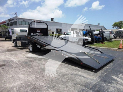 Isuzu NPRR rollback tow truck flatbed in a dealership lot with other equipment in background.