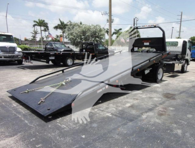 Isuzu NPRR rollback tow truck with a long flatbed in a dealership lot, white cab, metal black flatbed, warning decals, other trucks in background.