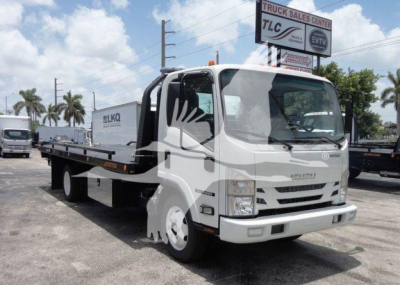 Isuzu NPR flatbed rollback truck in a dealership lot, front and left-side view with crane-style cab, warehouse background, blue sky.