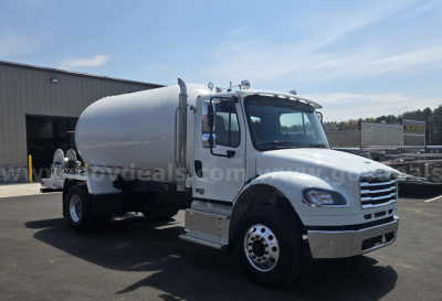 Freightliner M2 tank truck in a parking lot with white cab and large cylindrical tank mounted behind. Text on side windows and background not legible.