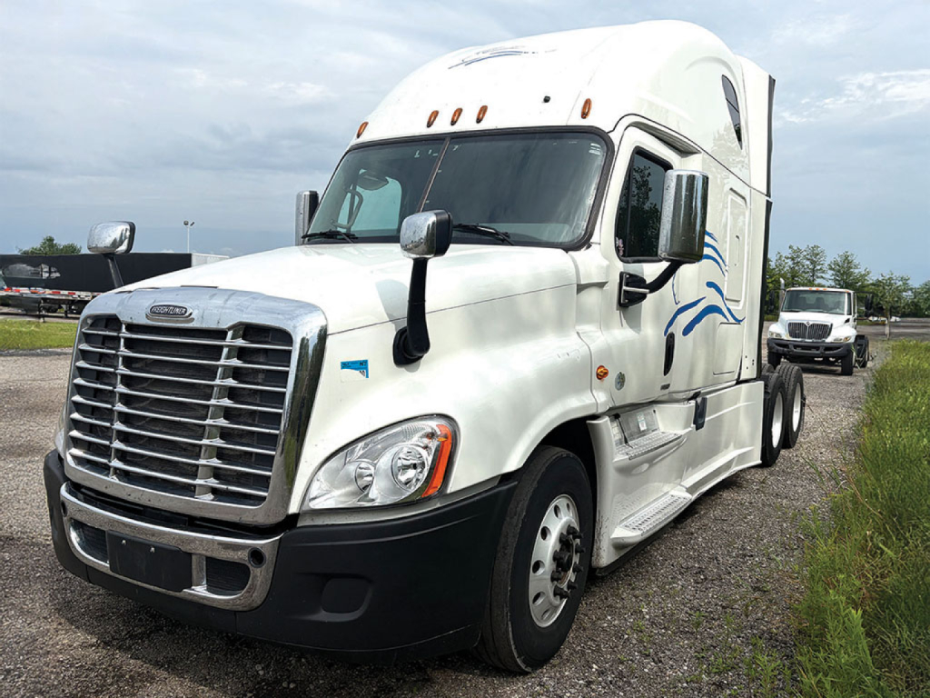 Truck cab of a white Freightliner Cascadia125 semi-truck with large grille, side mirrors, and blue decorative graphics; visible windows and front bumper