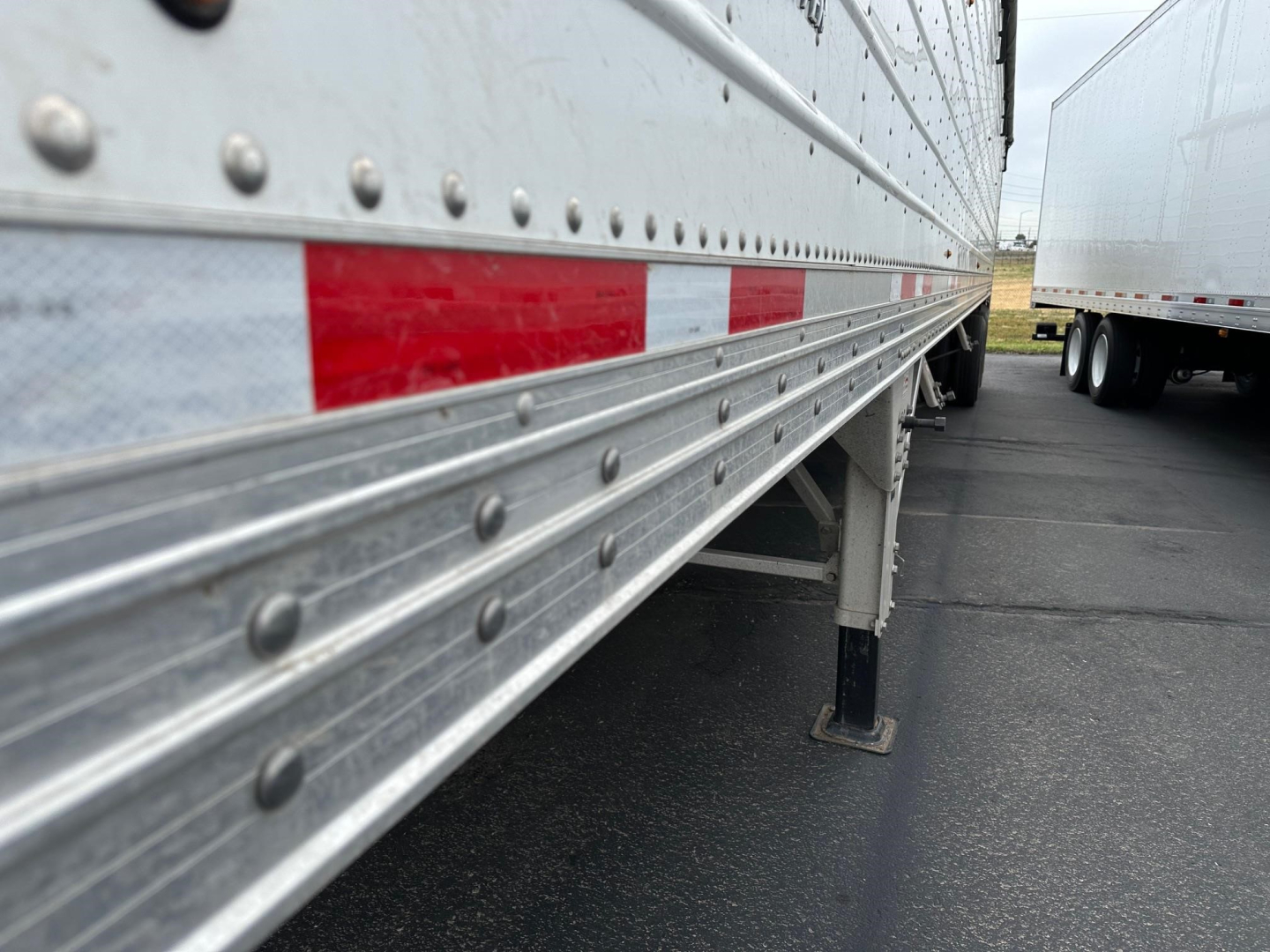 Trailer side view showing aluminum hopper trailer with riveted panels, red and white reflective tape, support leg, and adjacent trailer.