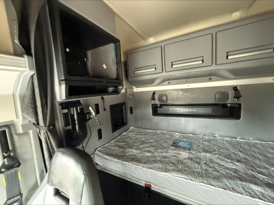 Truck cab bunk area of a 2020 Mack Anthem sleeper with storage cabinets and a folded-drawer overhead, visible window shade, and mattress wrapped in plastic.