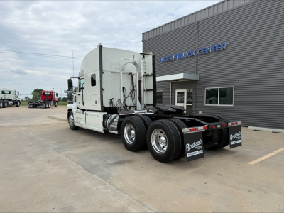 Truck tractor viewed from rear-left side showing white cab, three rear axles, fuel tanks, fifth wheel, and mud flaps. Buildings in background with blue sign.