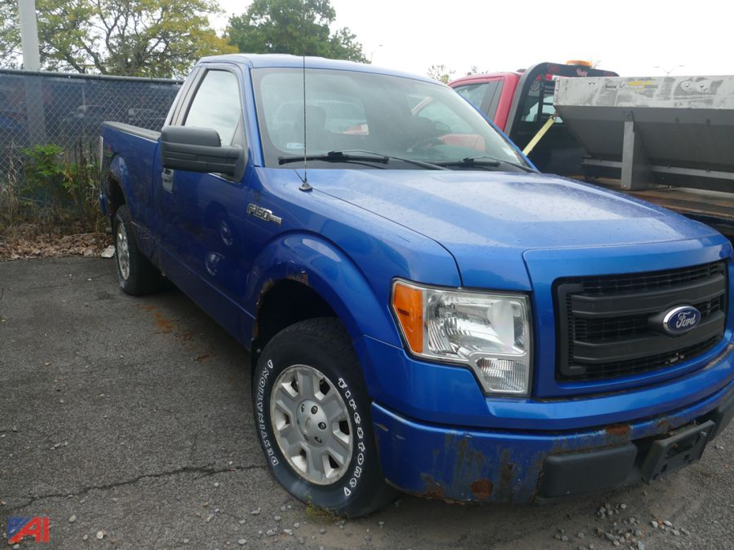 Blue Ford F-150 pickup truck, front three-quarter view, showing front bumper, grille, and right-side door; dented lower front bumper and wheel with steel rim; rain droplets on surface.