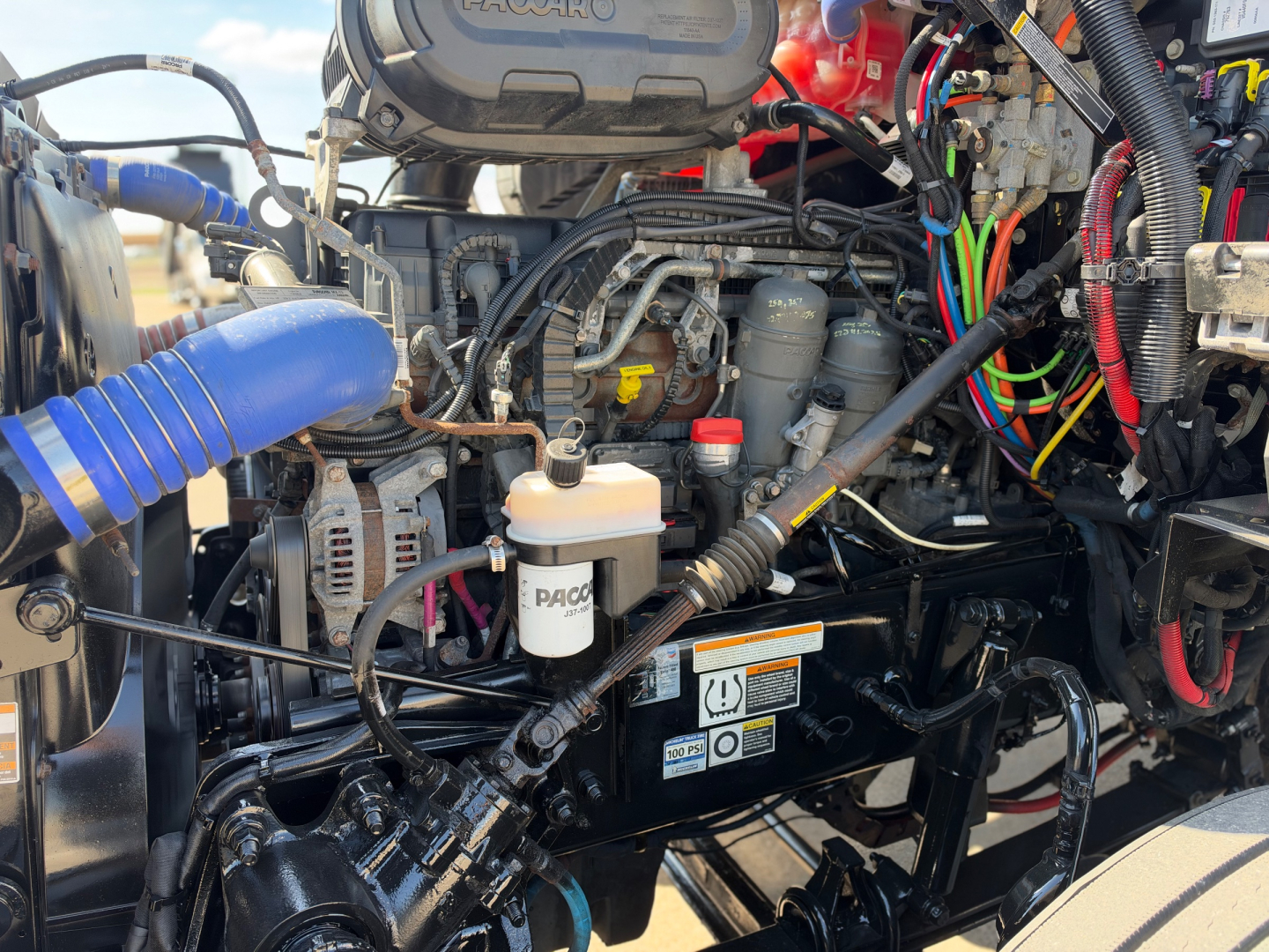 Peterbilt 579 engine bay with PACCAR engine and wiring harnesses, coolant reservoir, and various hoses visible.