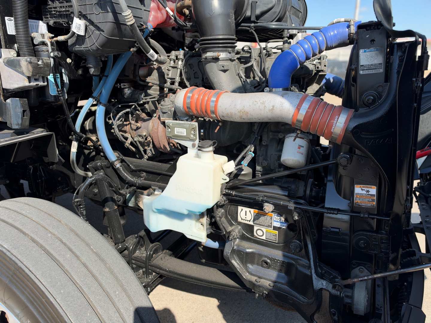 Peterbilt 579 engine bay with coolant reservoir, hoses, and components visible; blue silicone hose, white coolant reservoir, and orange-striped hose fittings in view.