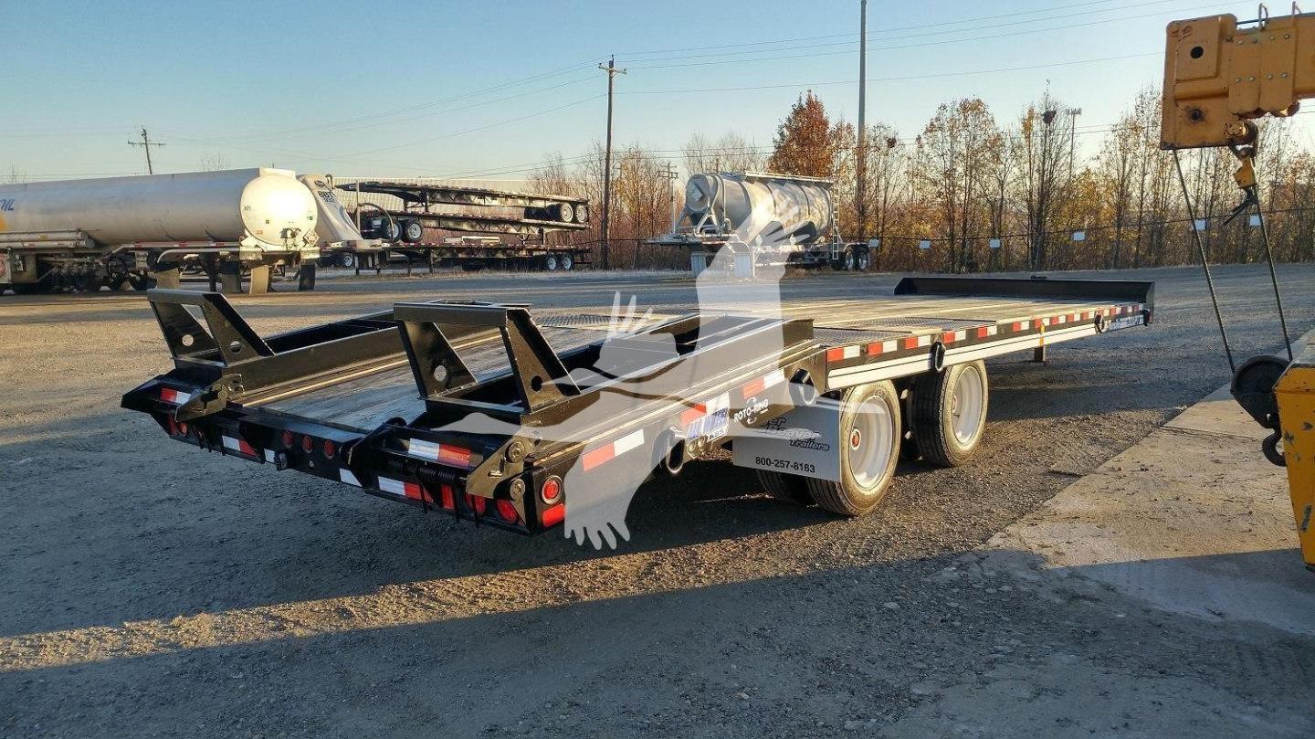 Flatbed trailer in an outdoor lot showing lower deck with wheel axles and ramps folded along the side; equipment and other trailers in background.