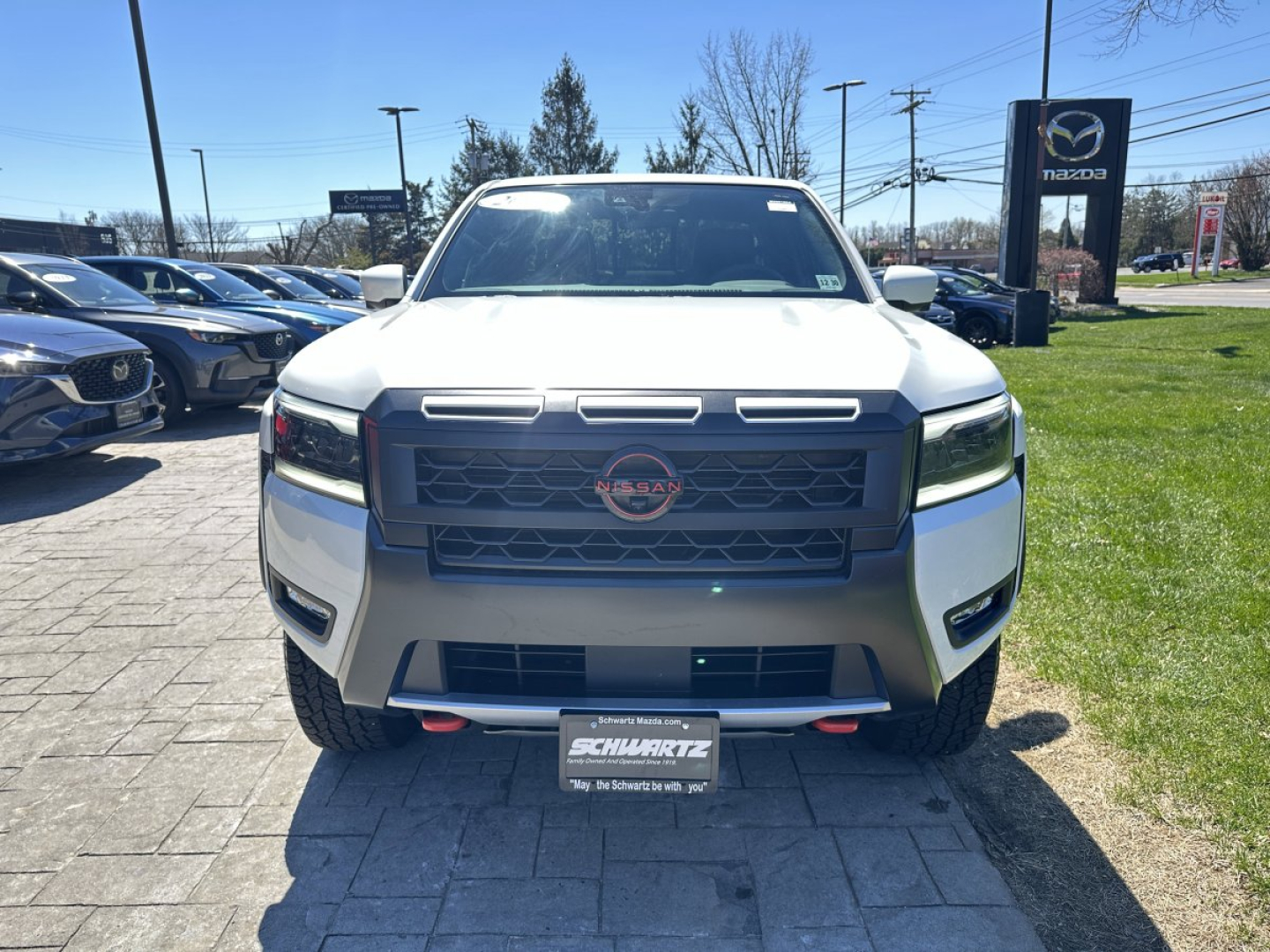 Front view of a white 2026 Nissan Frontier pickup truck in a dealership lot with Nissan badge on grille and black stock bumper; visible front license plate frame from Schwarcz.