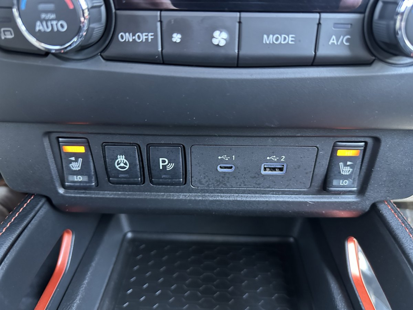 Nissan Frontier interior center console showing climate controls, auxiliary switches, and storage pocket; visible icons include heated seat, parking sensor, USB ports, and power button.