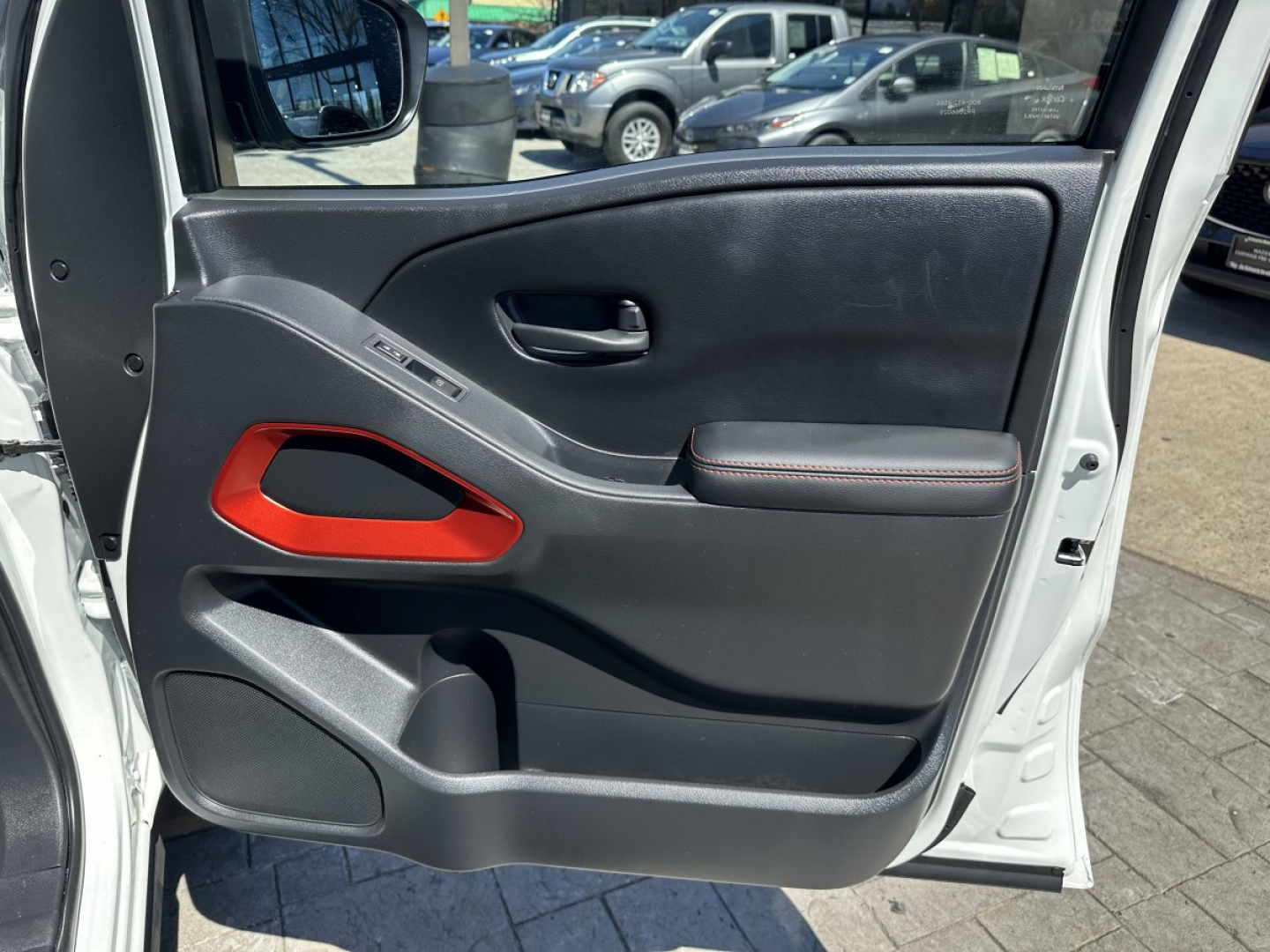 Vehicle interior door panel of a 2026 Nissan Frontier showing black plastic with red accent, door handle, window controls, storage pocket, and armrest.