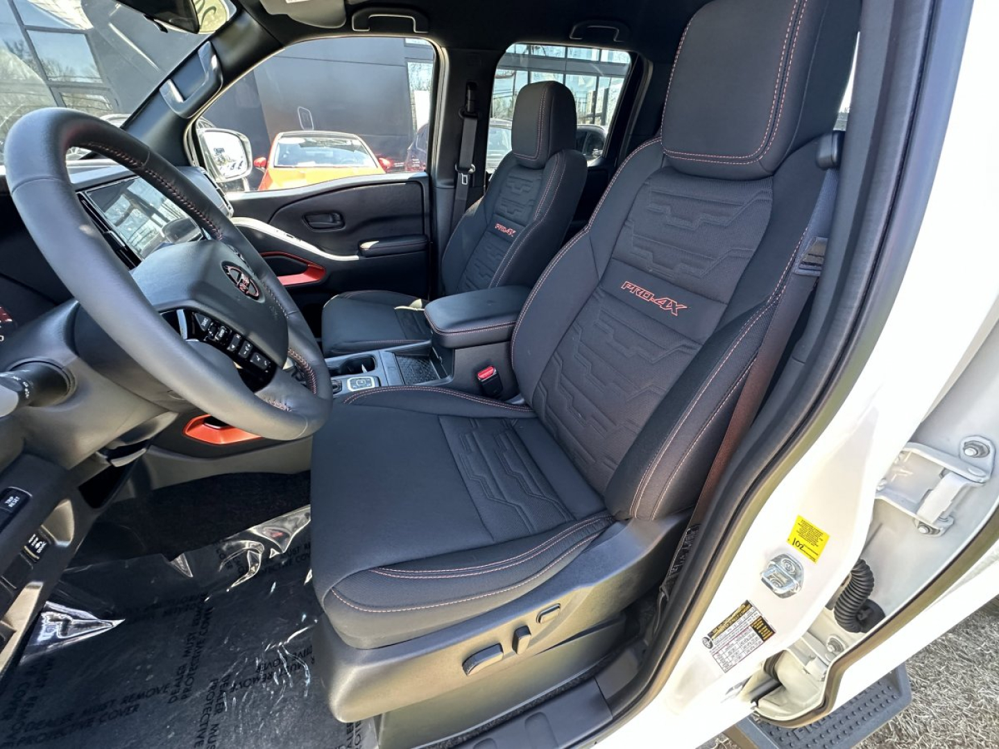 Interior view of a 2026 Nissan Frontier crew cab showing front seats and steering wheel with red stitching and 'Frontier' embroidered on seat, driver-side door open.
