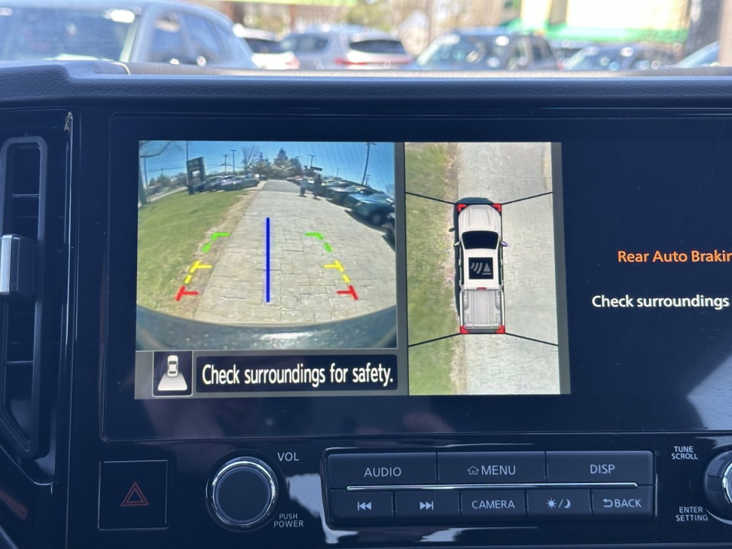 Vehicle infotainment screen showing rearview camera and top-down 360 view with guiding guidelines in yellow, green, and red. Message: Check surroundings for safety.