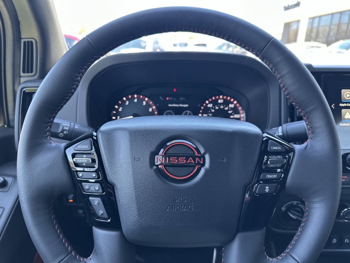 Inside view of a 2026 Nissan Frontier steering wheel with Nissan emblem and control buttons on both sides, dashboard gauges visible.