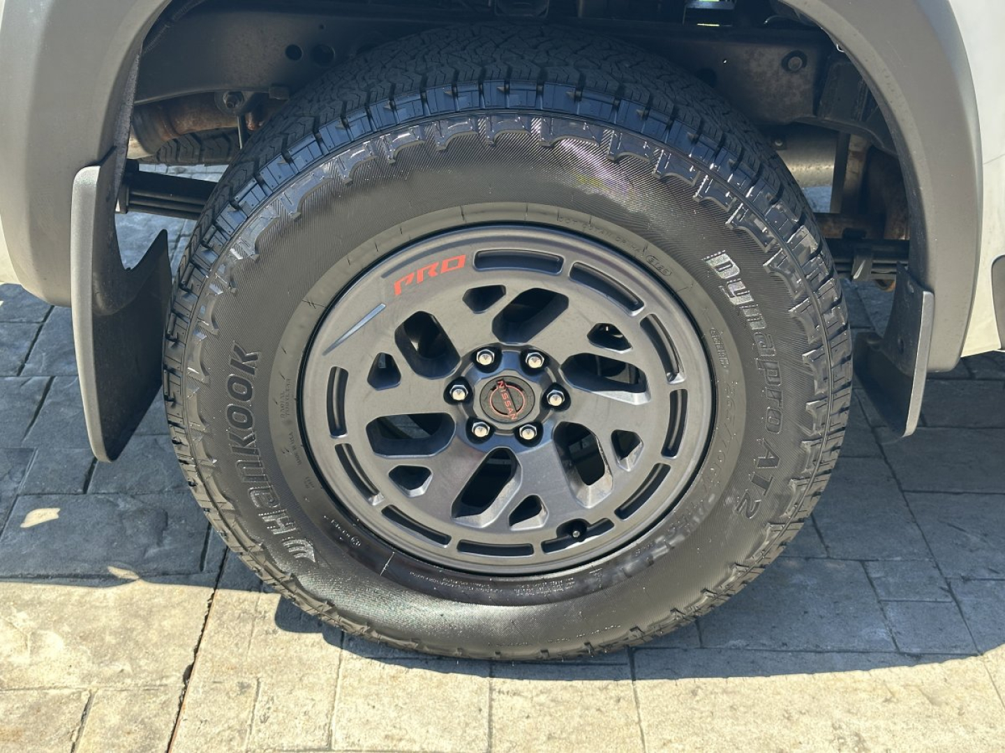 Wheel and tire of a Nissan Frontier pickup showing BFGoodrich All-Terrain T/A tire on a black alloy wheel with Nismo center cap and Pro-Comp styling; view focuses on front wheel well.