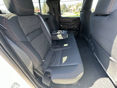 Back seat area of a 2026 Nissan Frontier showing dark cloth seats with orange stitching and center fold-down armrest. Rear doors and floor carpeting visible.