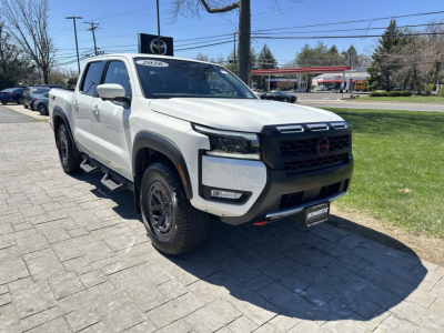 2026 Nissan Frontier pickup truck in white, front three-quarter view showing stock front grille and bumper, with dealership plate, side step, black wheels, and label on windshield.