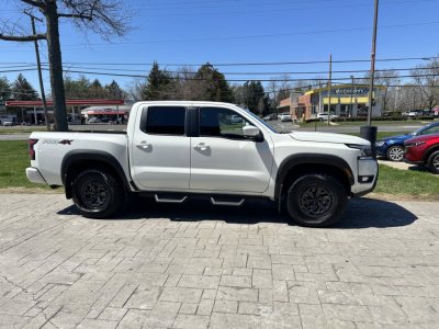 Side profile of a white 2026 Nissan Frontier pickup truck in a dealership lot with crew cab and black alloy wheels.