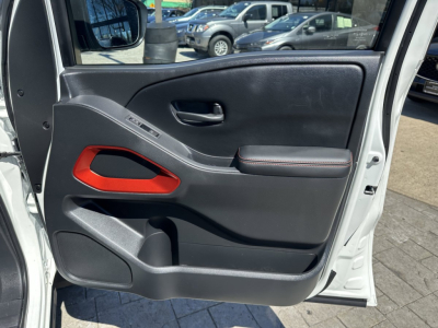Vehicle interior door panel of a 2026 Nissan Frontier showing black plastic with red accent, door handle, window controls, storage pocket, and armrest.