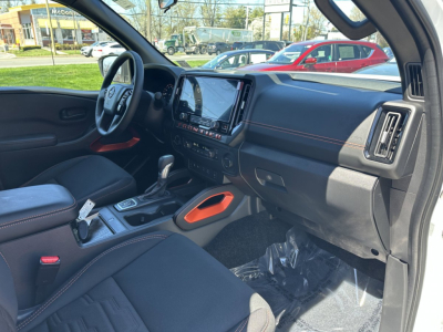 Nissan Frontier interior view from the passenger side showing black cloth seats, center console with touchscreen display, steering wheel, and orange accents near the cup holder area.