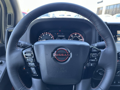 Inside view of a 2026 Nissan Frontier steering wheel with Nissan emblem and control buttons on both sides, dashboard gauges visible.