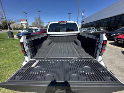 Truck bed of a white 2026 Nissan Frontier with open tailgate, showing cargo bed liner and wheel wells, dealership lot in background.