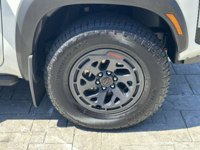 Nissan Frontier wheel and tire with black alloy rim mounted on front fender area; tire sidewall shows 'TOURING' style tread and 'Goodyear Wrangler' branding; circular center cap with emblem.