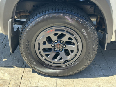 Wheel and tire of a Nissan Frontier pickup showing BFGoodrich All-Terrain T/A tire on a black alloy wheel with Nismo center cap and Pro-Comp styling; view focuses on front wheel well.